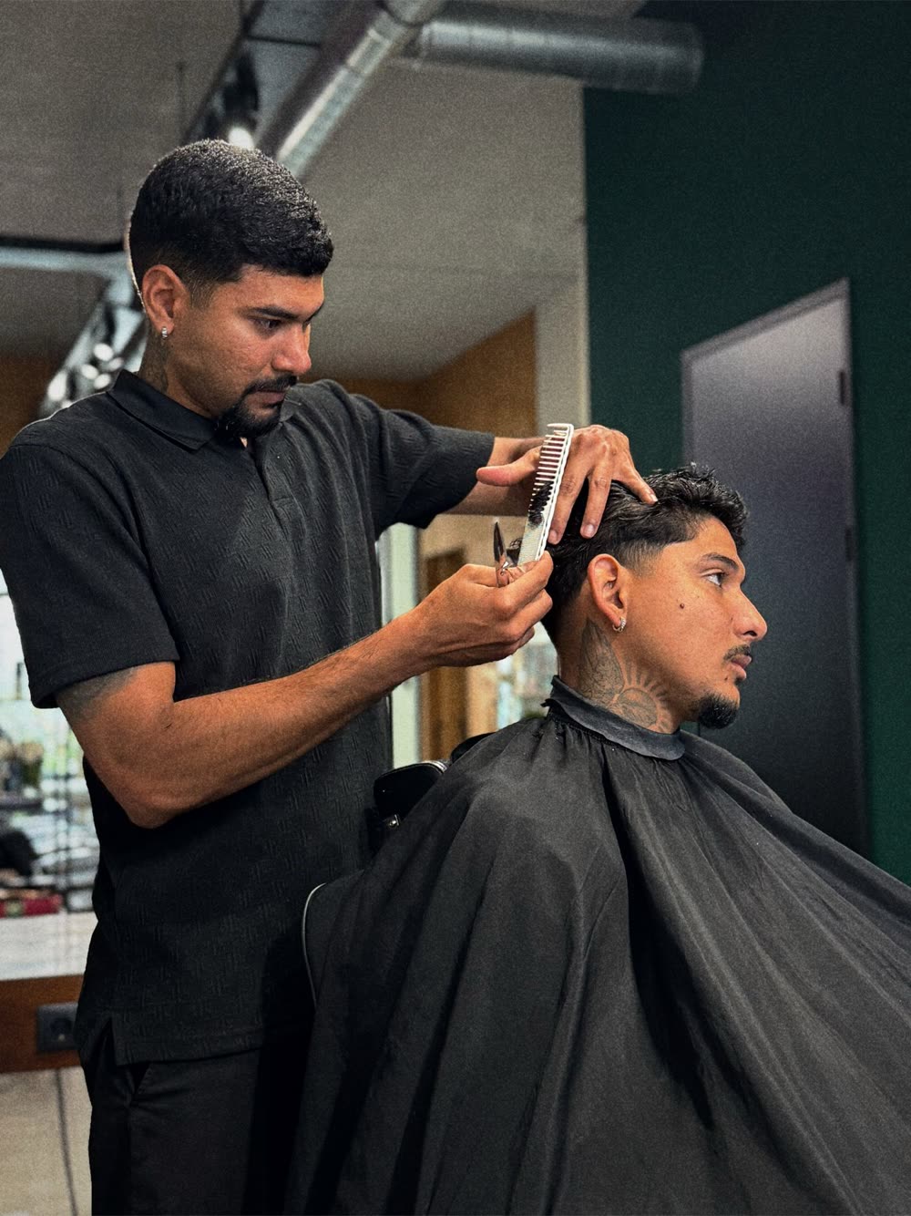Johnathan cortando o cabelo de um cliente na tesoura e no pente, ao lado da parede verde