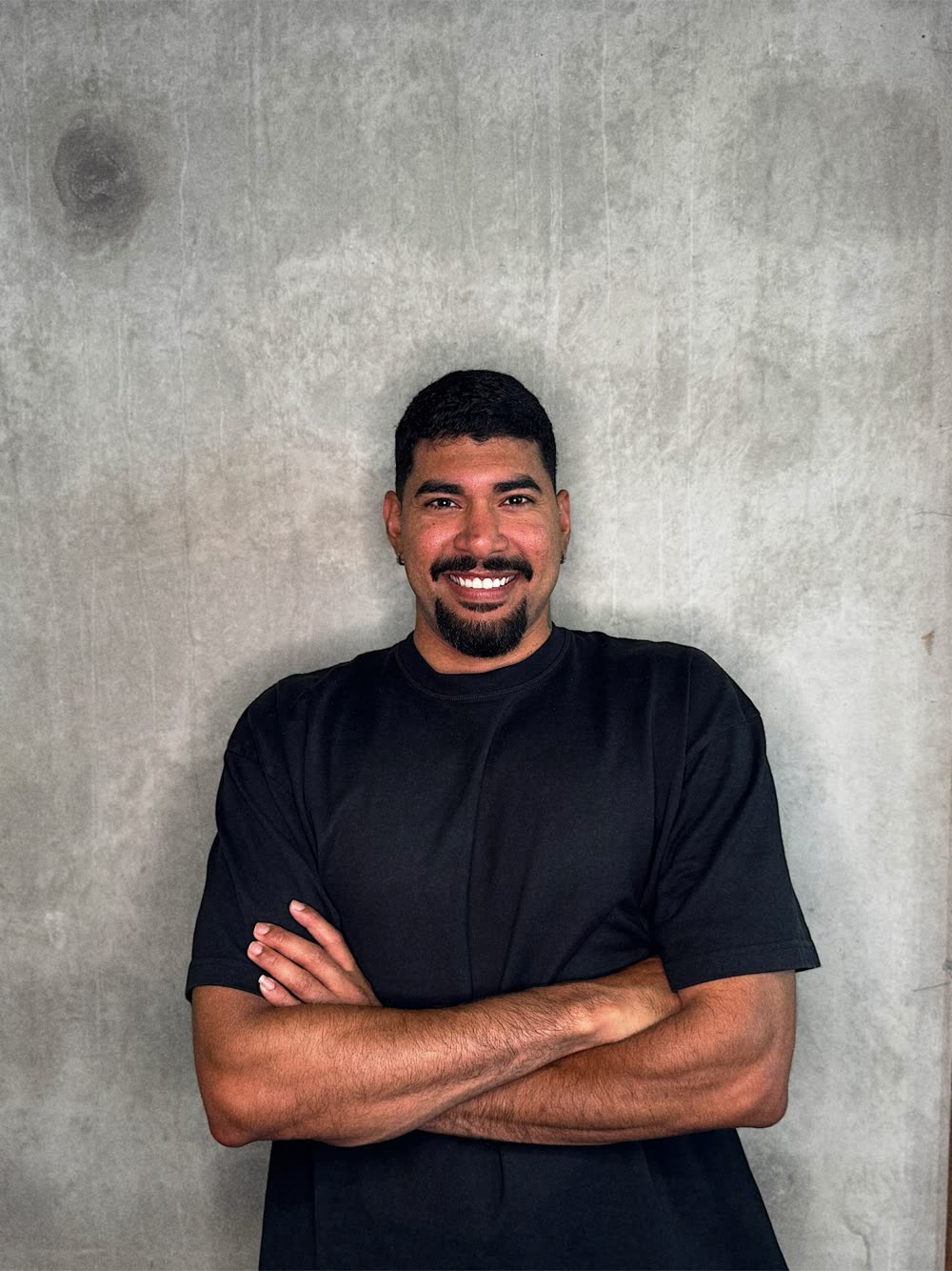 Johnathan, de camiseta preta e braços cruzados, sorrindo diante de uma parede de concreto